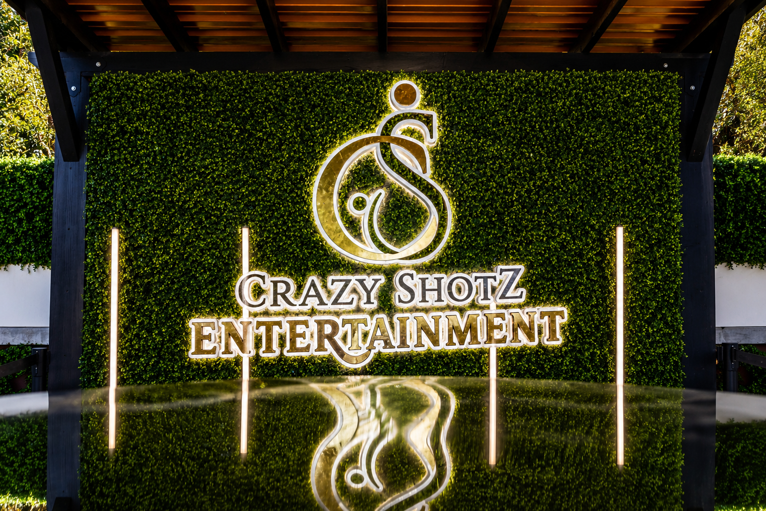 Illuminated Crazy Shotz Entertainment gold logo on a lush hedge wall with reflective floor